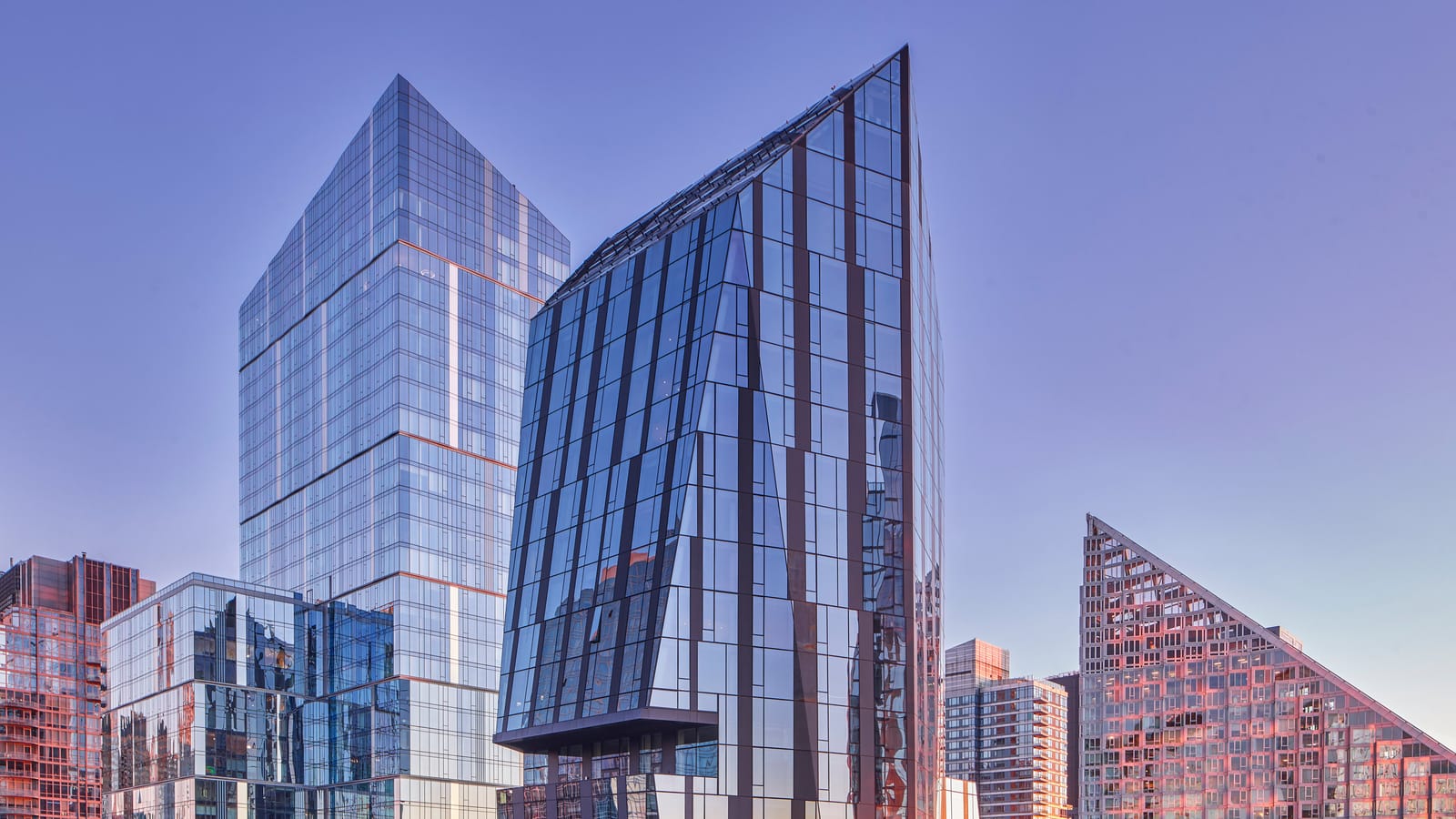 The top of a faceted glass apartment tower with a sharply sloped, pointed crown and a dark ledge jutting out near its base, against a clear dusk sky, with a taller square glass tower behind it at left and a red-lit, triangle-shaped building at right
