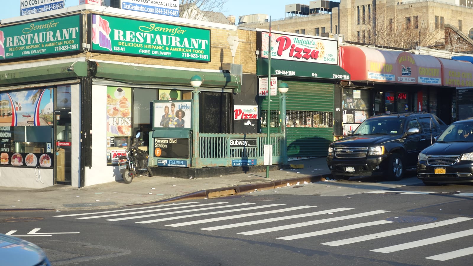 A green railed subway stair on a street corner with a sign reading Parsons Blvd Station and an orange F symbol, beside a restaurant and a pizza shop, with parked cars at right and a crosswalk in front