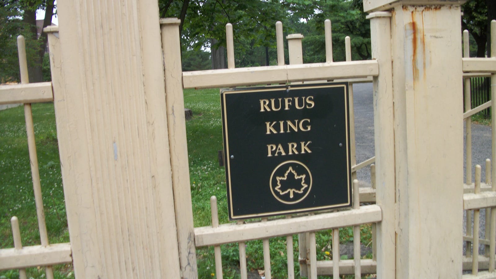 A black sign with gold letters reading Rufus King Park and the Parks Department leaf, on a cream-painted iron gate between two pillars, with grass, trees and a paved path behind