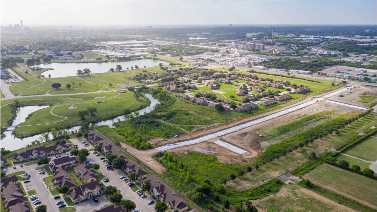 Aerial photograph of a graded strip of land with a new concrete street, beside a park with a lake and rows of brown-roofed apartment buildings, with a skyline on the horizon
