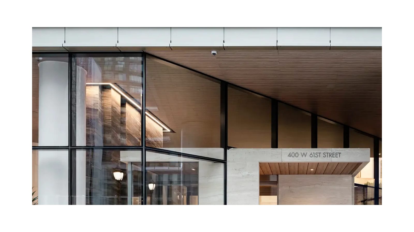 A glass and stone building entrance under a wood-slatted canopy, with the street number 400 W 61st Street carved on a pale stone frame over the door and a tall glass lobby wall at left