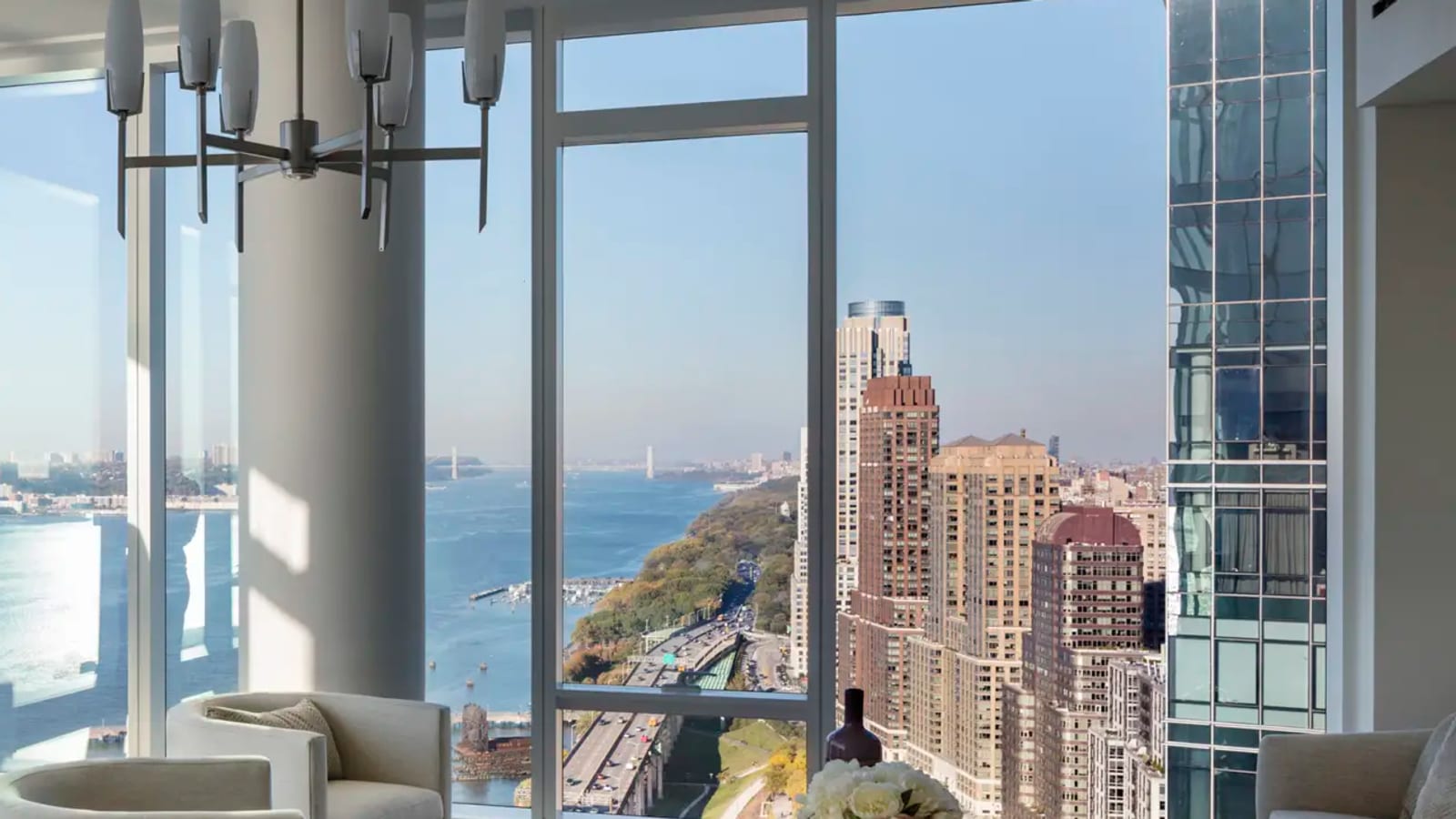 A corner living room with floor-to-ceiling windows looking north over the Hudson River, a highway and a row of tall brick apartment towers, with white armchairs, a sofa and a glass coffee table under a chandelier