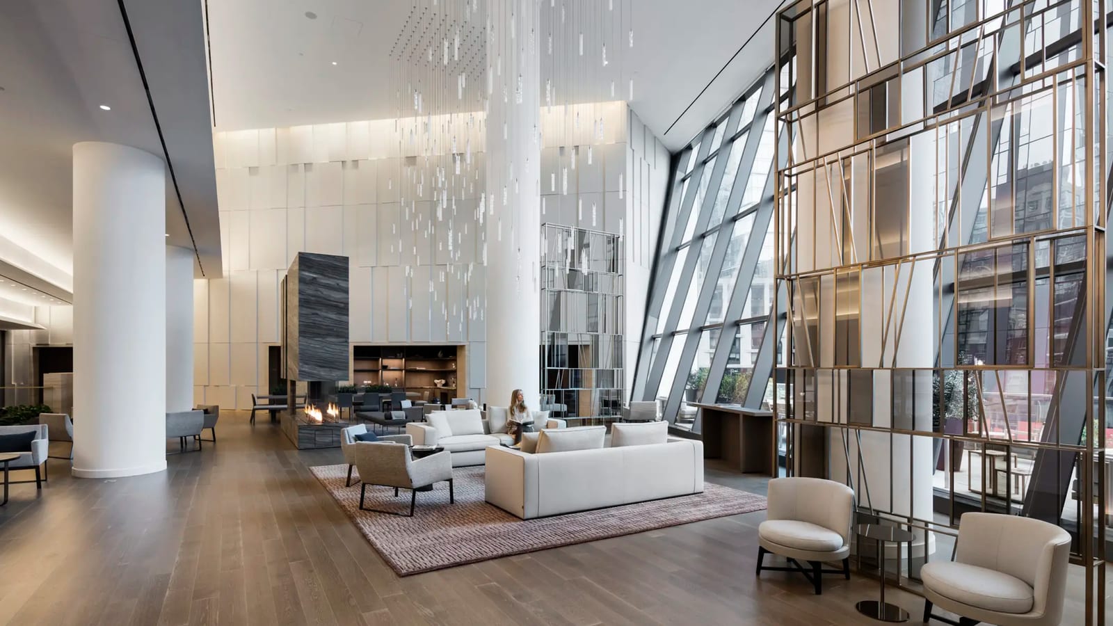 A tall, bright lobby with white columns, a light fixture of hanging glass rods, a stone fireplace, white sofas on a rug and a bronze lattice screen in front of slanted glass walls