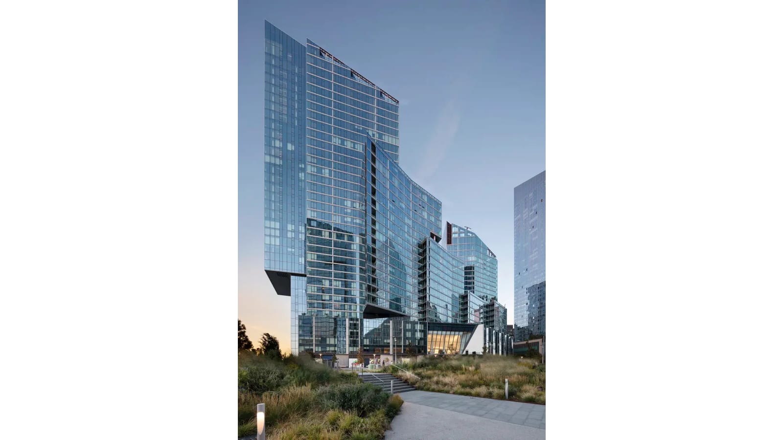 A glass tower rising in two stepped blocks over a lit glass lobby at dusk, seen from a park path lined with tall grasses and low light posts
