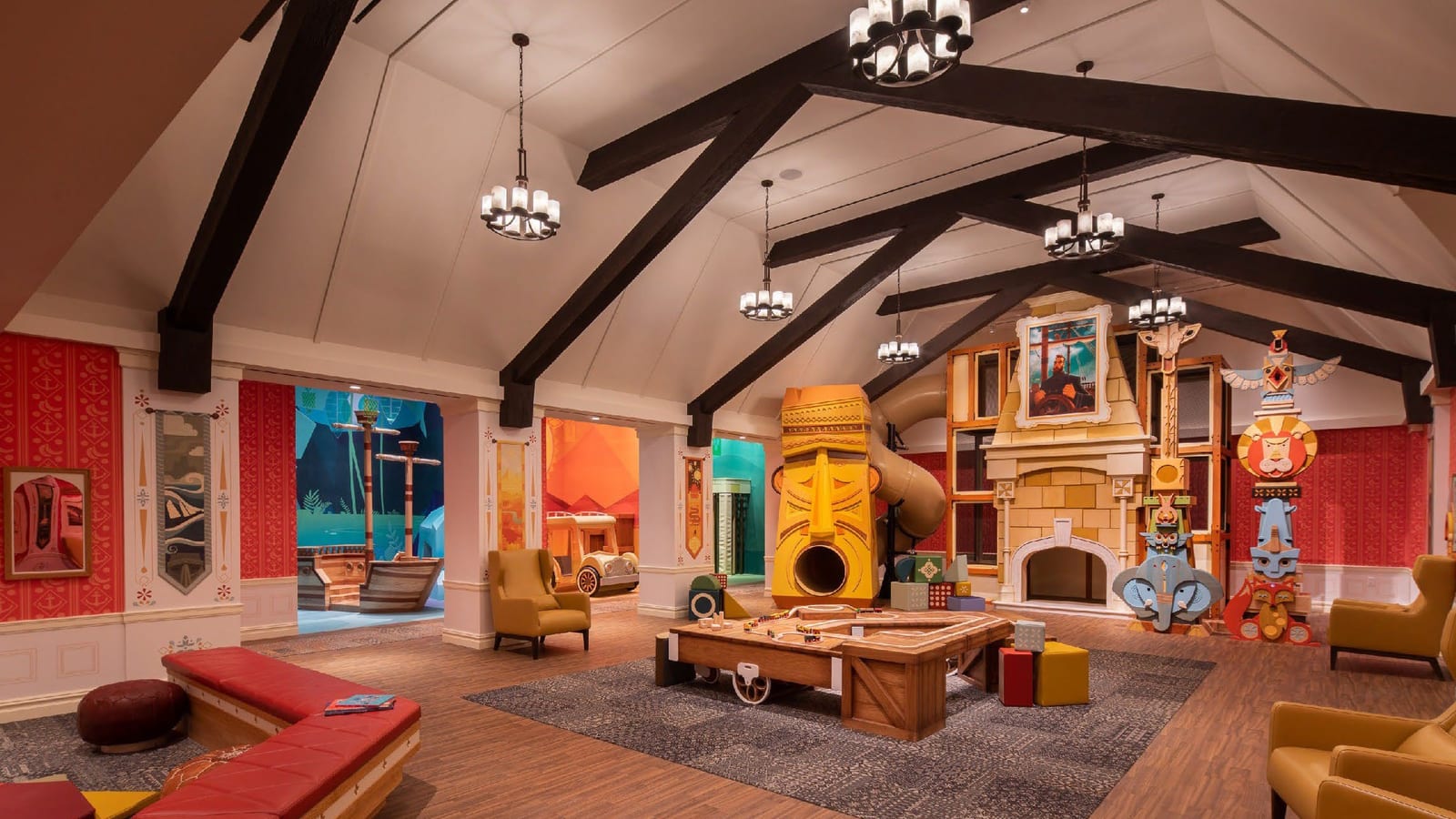 A large children's playroom under dark wooden roof beams, with a yellow tiki-style slide tower, a toy train table, a play fireplace, painted animal totems and red benches