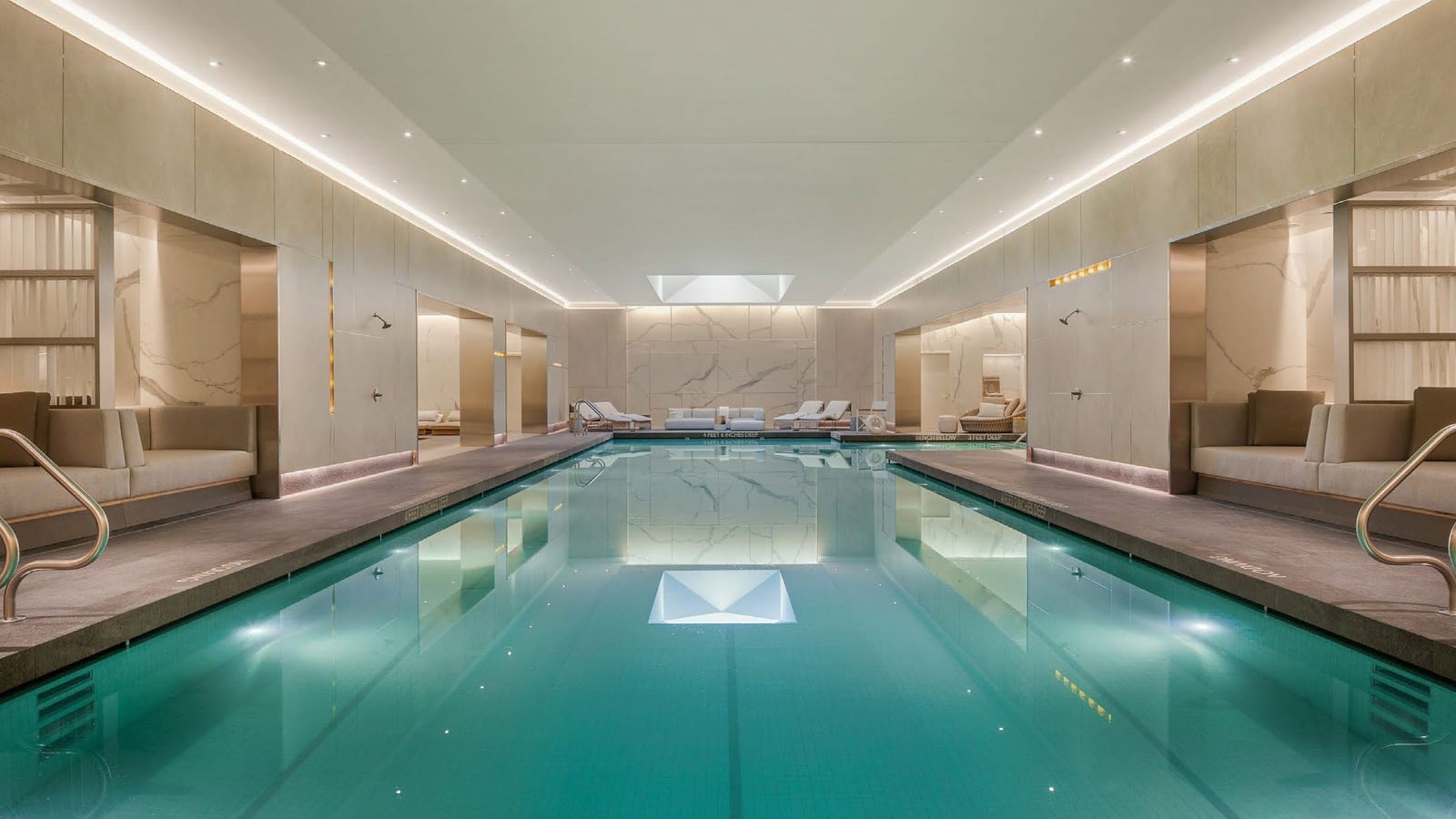 A long indoor lap pool with pale marble walls, cove lighting along the ceiling, steel ladders at the near end and lounge chairs at the far end