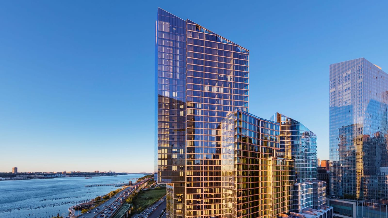 A tall glass apartment tower with stepped lower wings glowing gold in low sun, beside a wide highway and the Hudson River under a clear blue sky, with another glass tower at the right edge