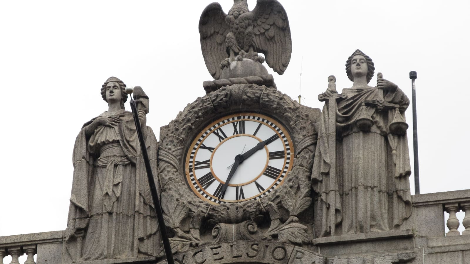 A stone clock with Roman numerals set in a carved wreath, an eagle above it and a robed stone figure on each side, with the word Excelsior below