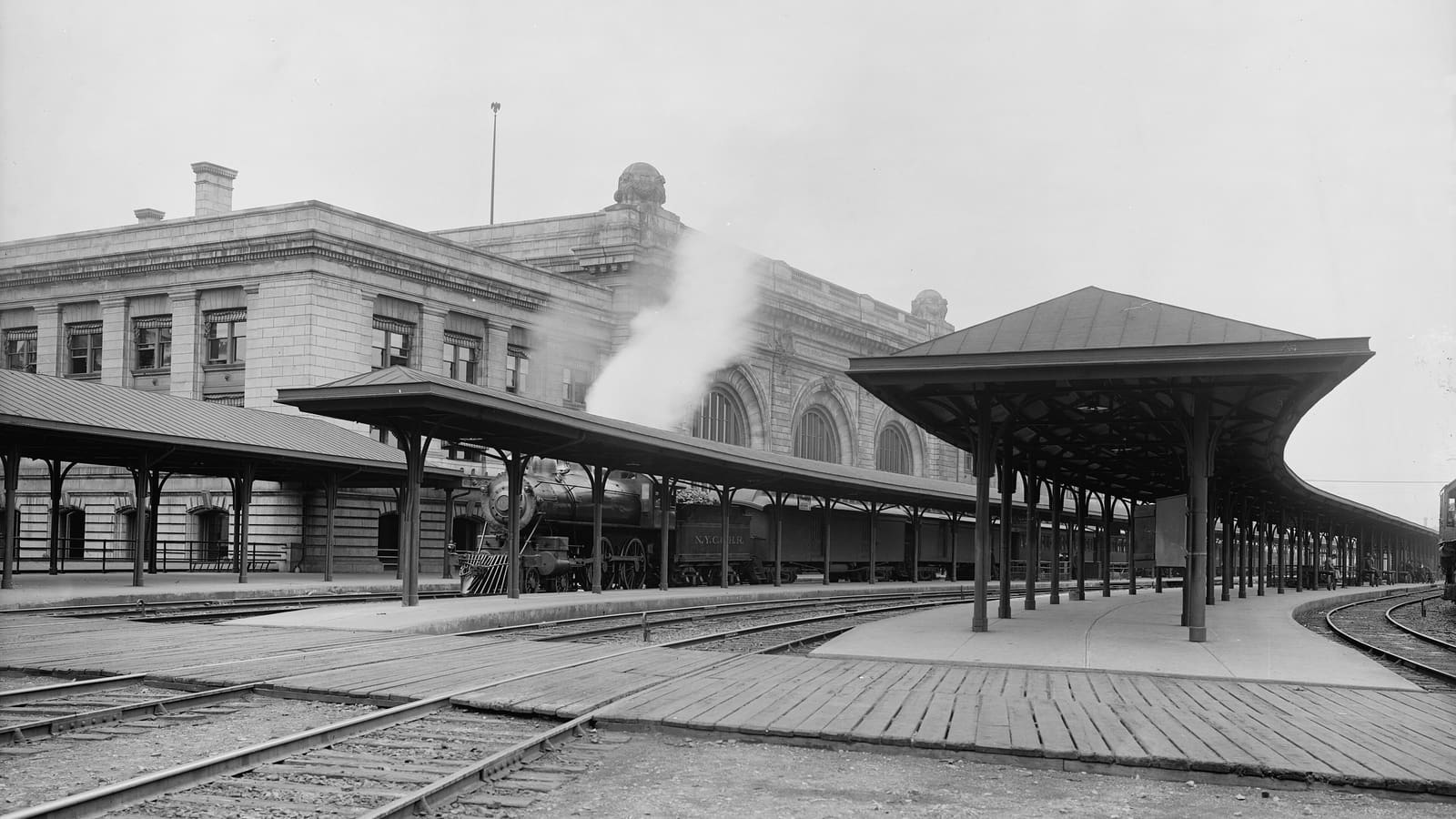 Black-and-white photograph of covered train platforms and tracks behind a stone station building, with steam rising from a locomotive
