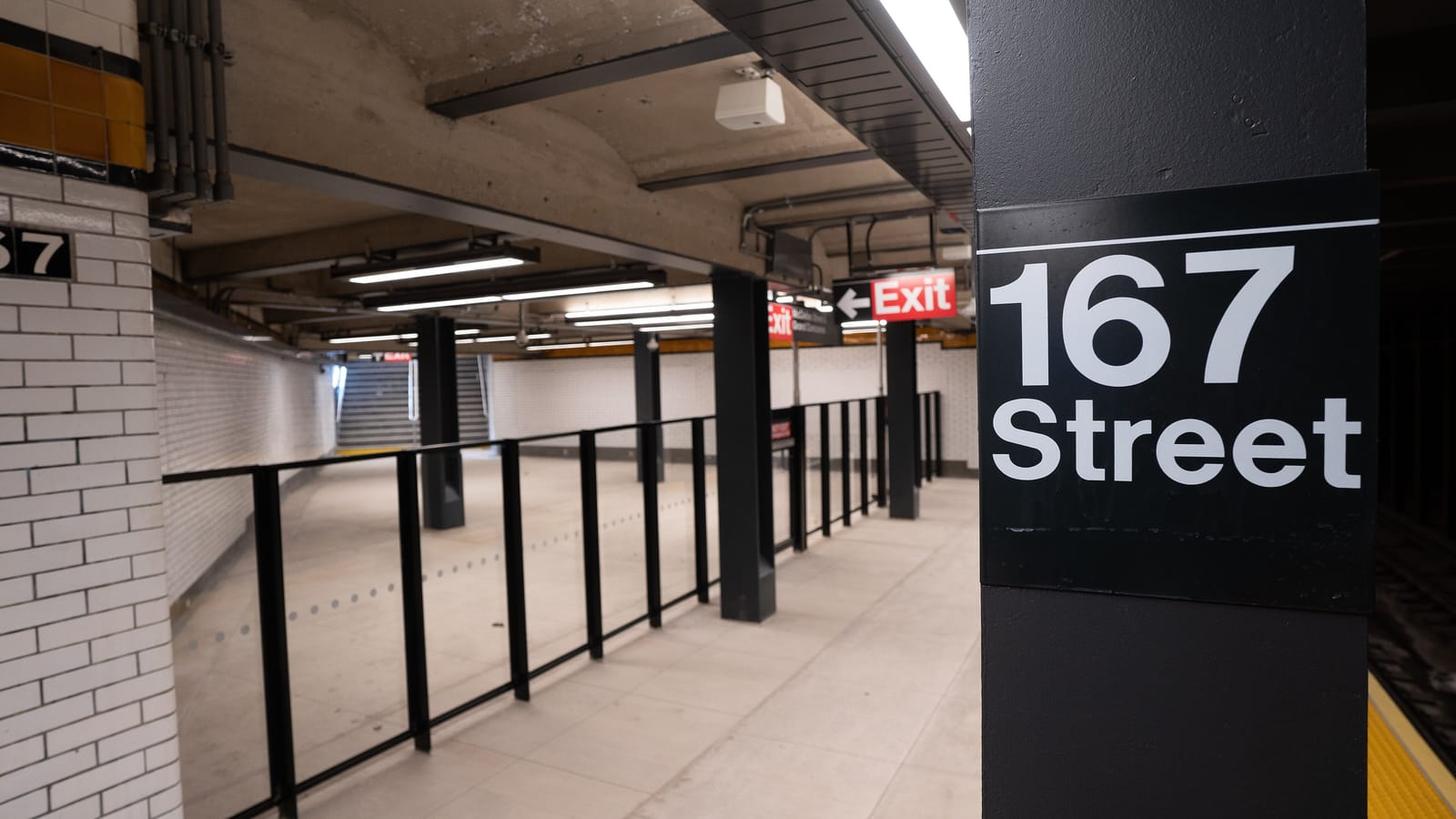 A subway platform with a black column carrying a white 167 Street sign, black railings, white tile walls, an orange tile band and red Exit signs over a stairway