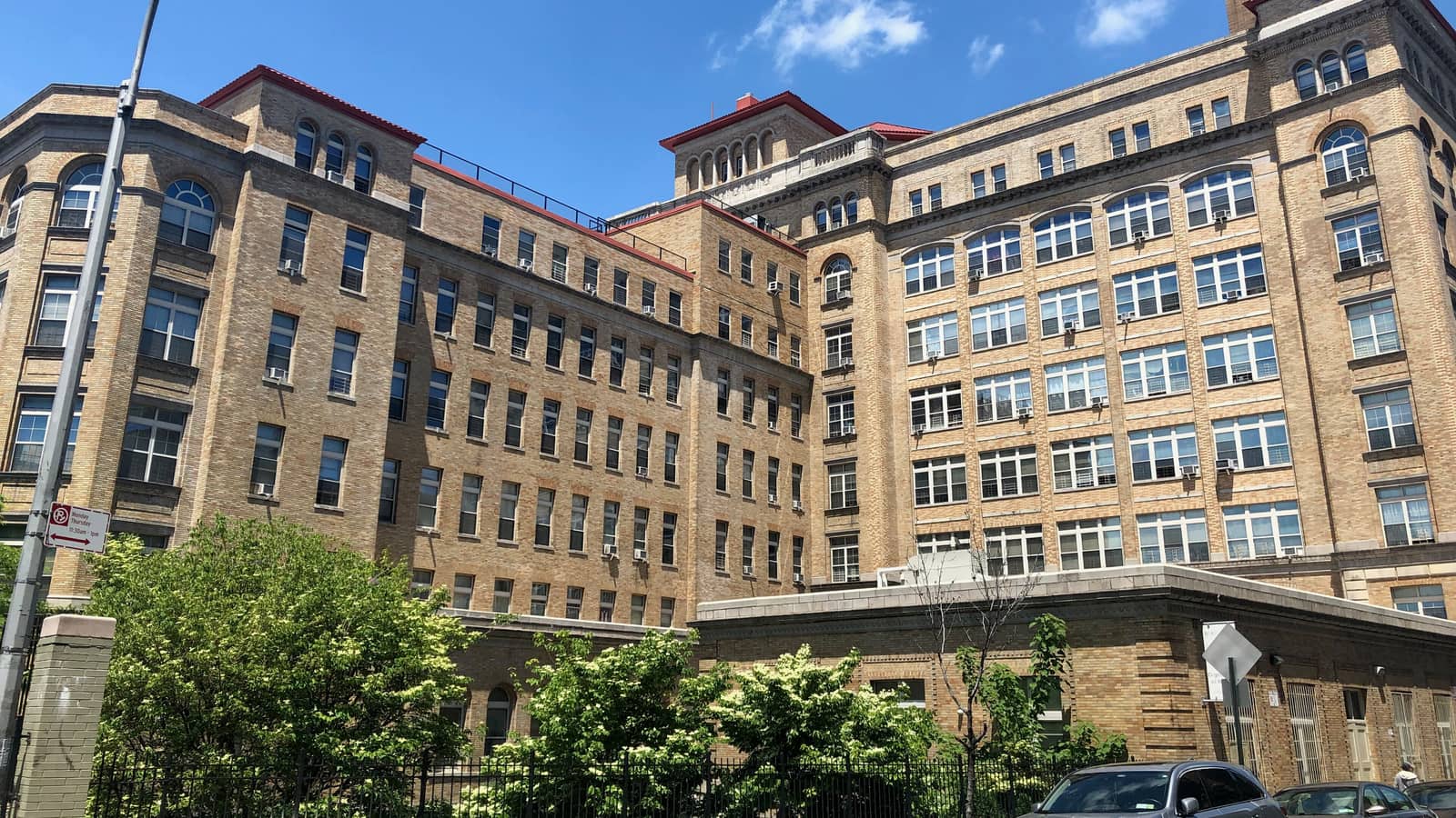 The long tan brick wings of the building around an open courtyard, seen over a black iron fence and flowering shrubs, with a low one-story wing at right and parked cars along the curb