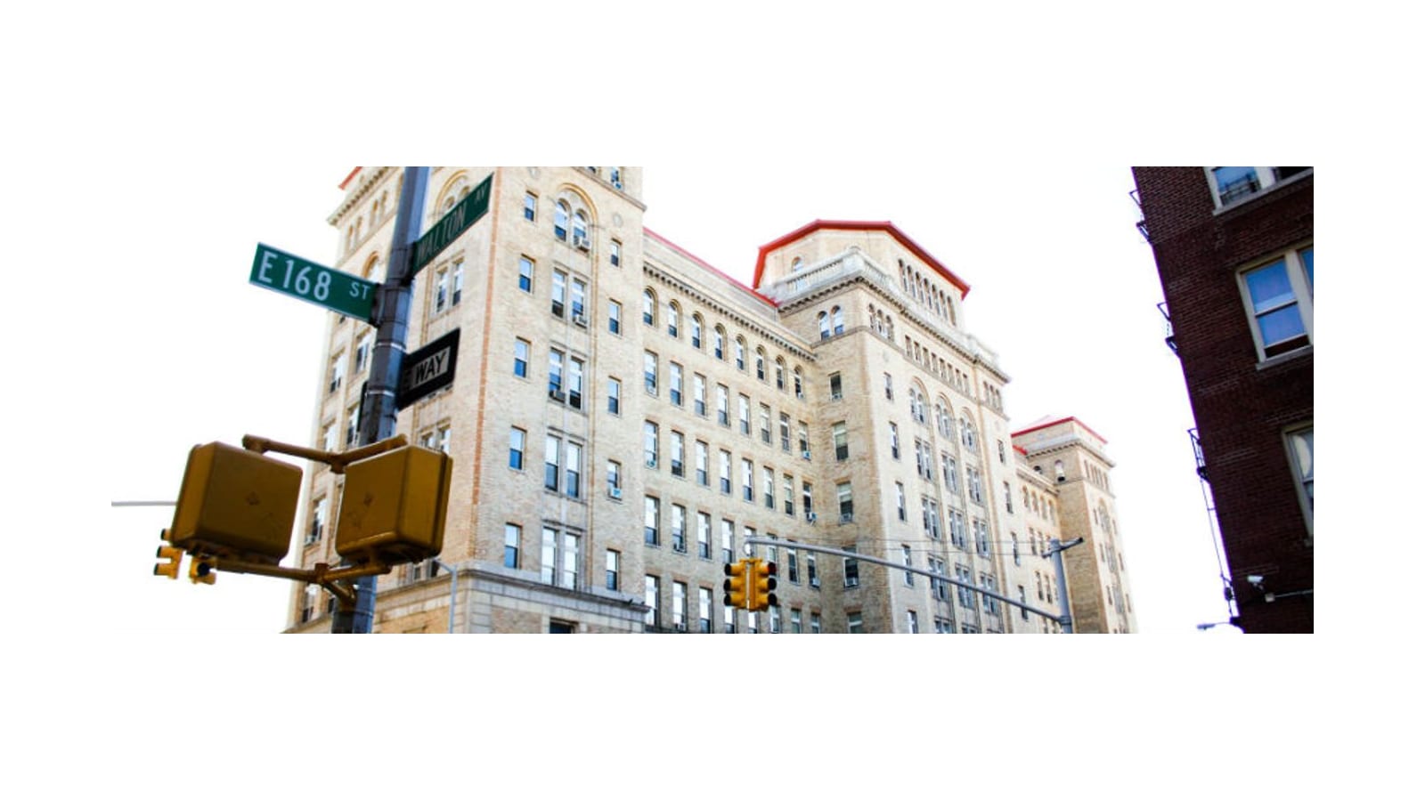 A tan brick building with red roof trim seen from below, behind a pole with green street signs for East 168th Street and Walton Avenue and yellow traffic lights
