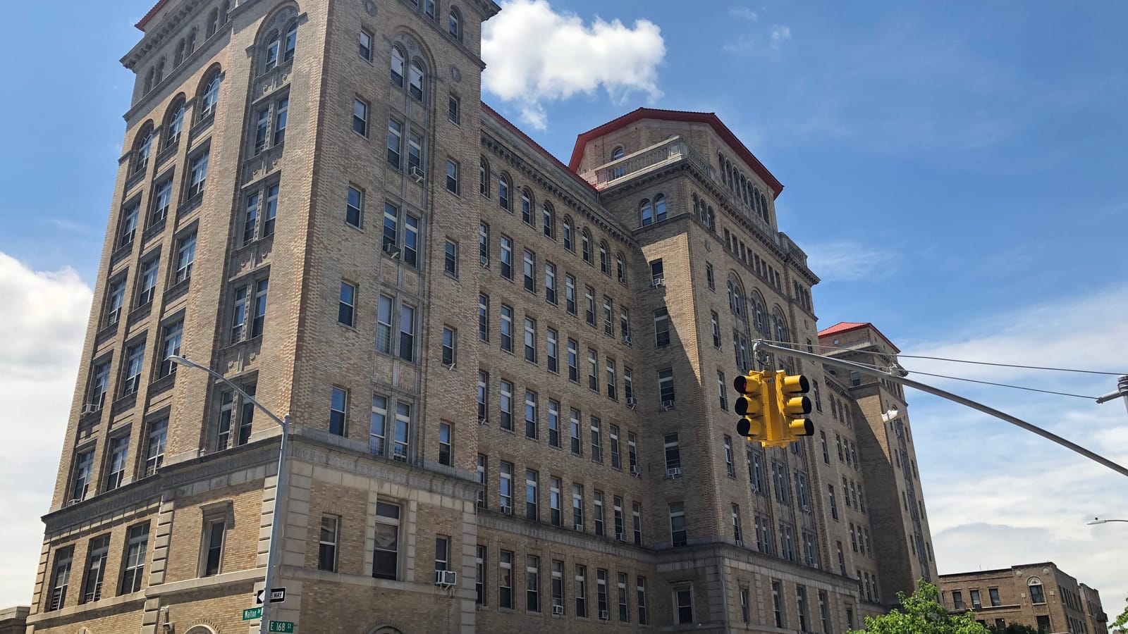 A tall tan brick corner tower with arched upper windows and a stone base, with street signs for Walton Avenue and East 168th Street on a pole and a yellow traffic light at right