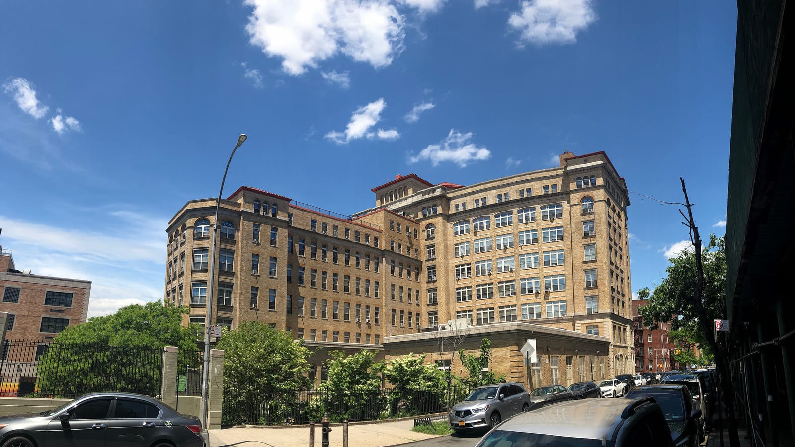 A ten-story tan brick building with red-tiled tower roofs and rows of arched windows, behind a fenced garden of shrubs and trees, with cars parked along the street under a blue sky