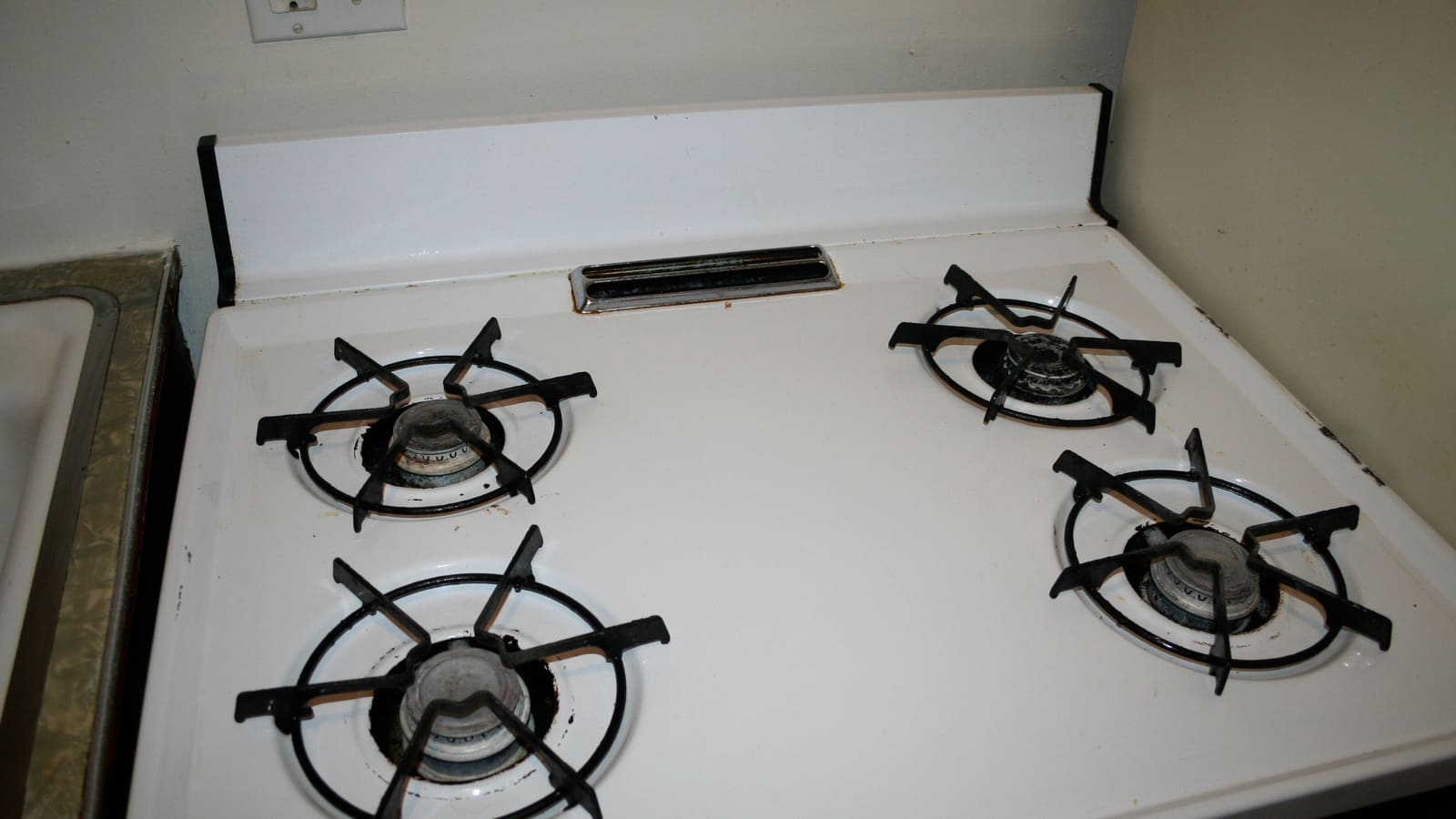The top of an old white kitchen stove with four gas burners and black grates, beside a sink