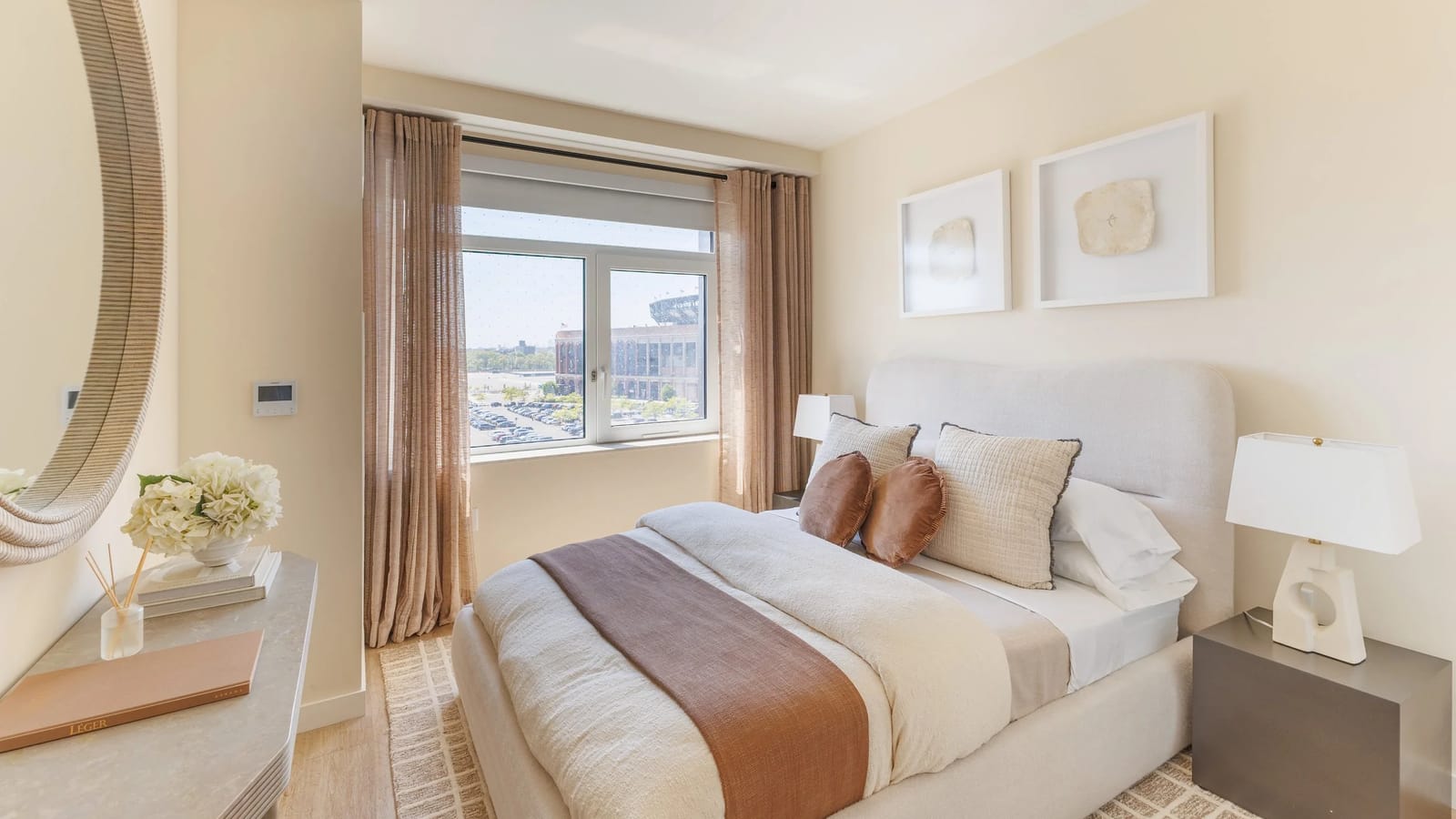 Photograph of a furnished bedroom with a bed, two bedside lamps, framed art and tan curtains beside a two-pane window that looks across a parking lot to a brick stadium, with a small control panel on the wall at the left