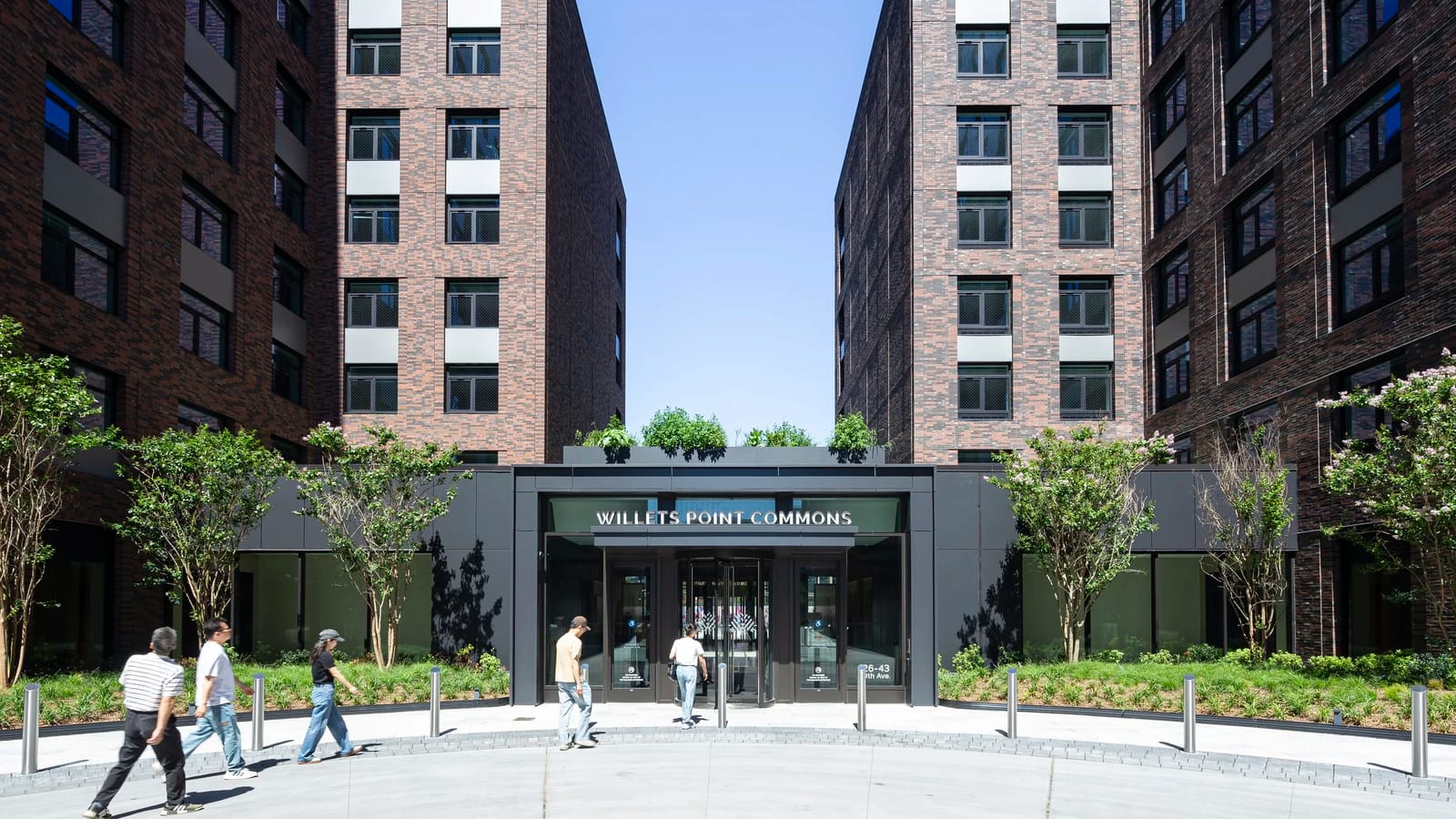 Photograph of a one-story dark metal entrance pavilion lettered WILLETS POINT COMMONS, with a revolving door and glass doors, set between two brick apartment wings with a strip of sky between them, flowering trees and planted beds on both sides, steel bollards along a curved driveway and five people walking past
