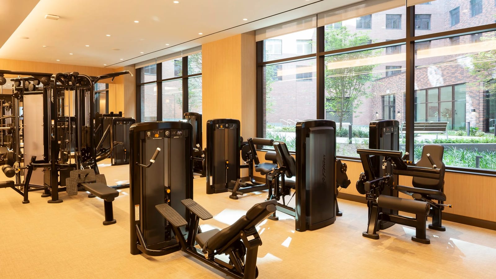 Photograph of a fitness room with black weight machines and a cable station on a pale floor, wood-panelled walls and tall windows looking onto a planted courtyard and brick apartment walls
