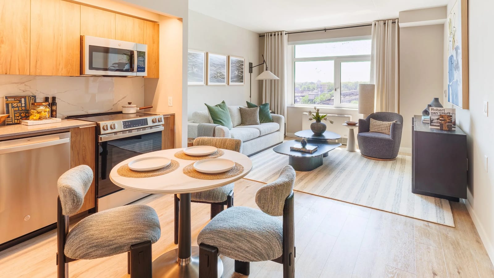 Photograph of a furnished apartment with a kitchen run at the left holding wood-tone cabinets, a stainless-steel dishwasher, a smooth-top electric range and a microwave, a round dining table with three chairs, and a living area with a sofa, an armchair, a striped rug and a wide window with a white unit below it