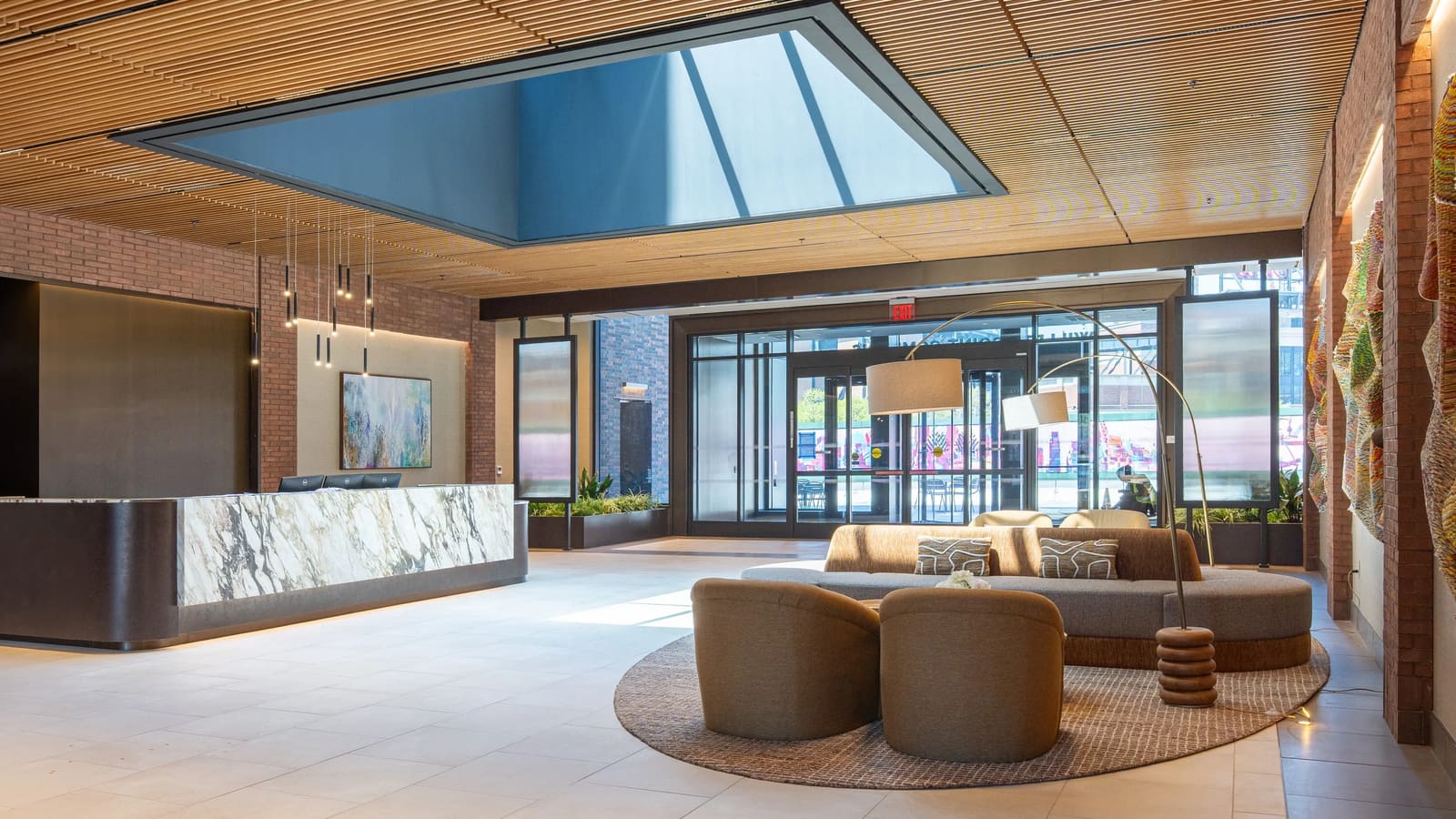 Photograph of a lobby with a slatted wood ceiling around a large skylight, a marble-fronted reception desk at the left, a round rug with curved sofas and armchairs, a floor lamp, brick walls and glass entrance doors opening to the street