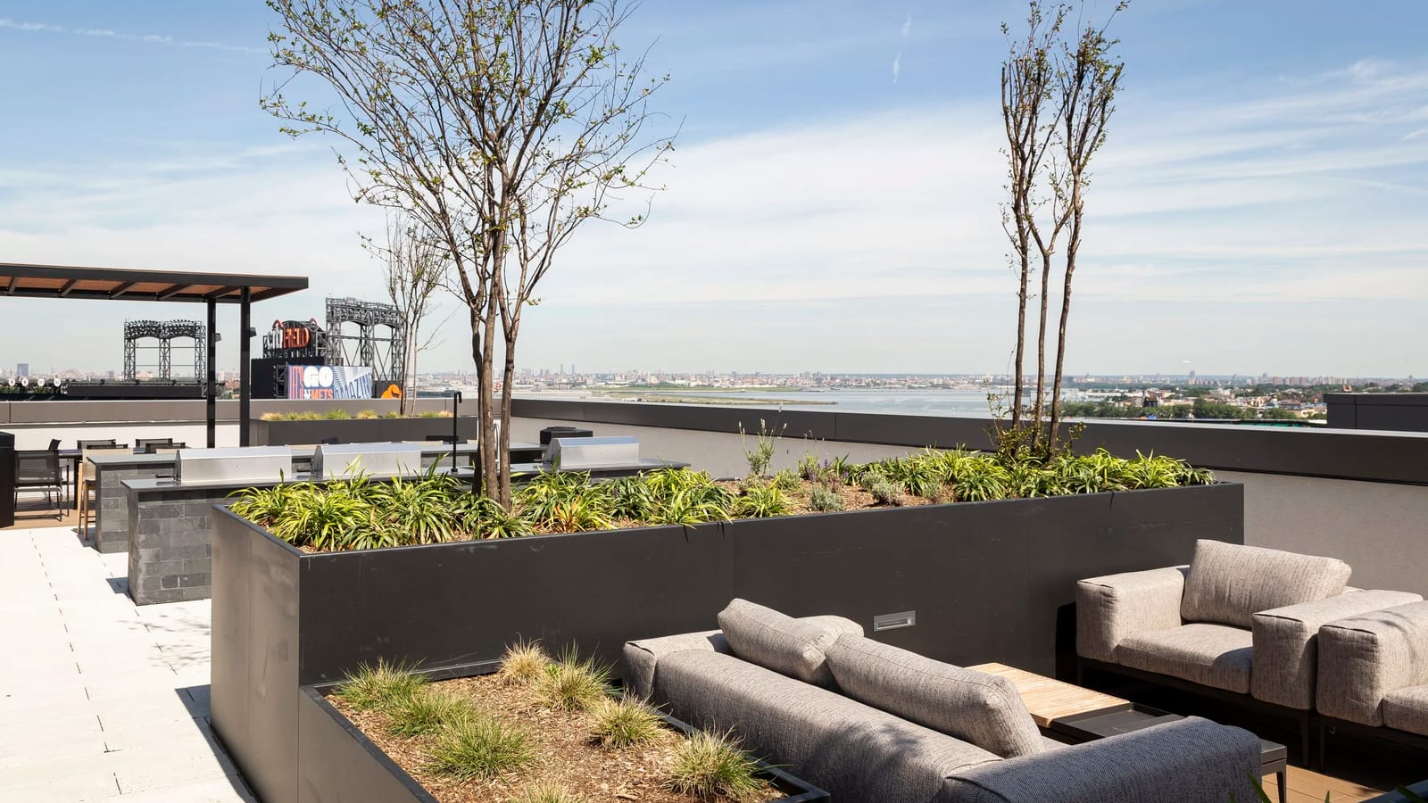 Photograph of a roof terrace with dark planters holding grasses and young trees, grey outdoor sofas, a row of stainless-steel grills on stone counters and a pergola, with stadium light towers and a GO METS sign at the left and a bay and low skyline in the distance