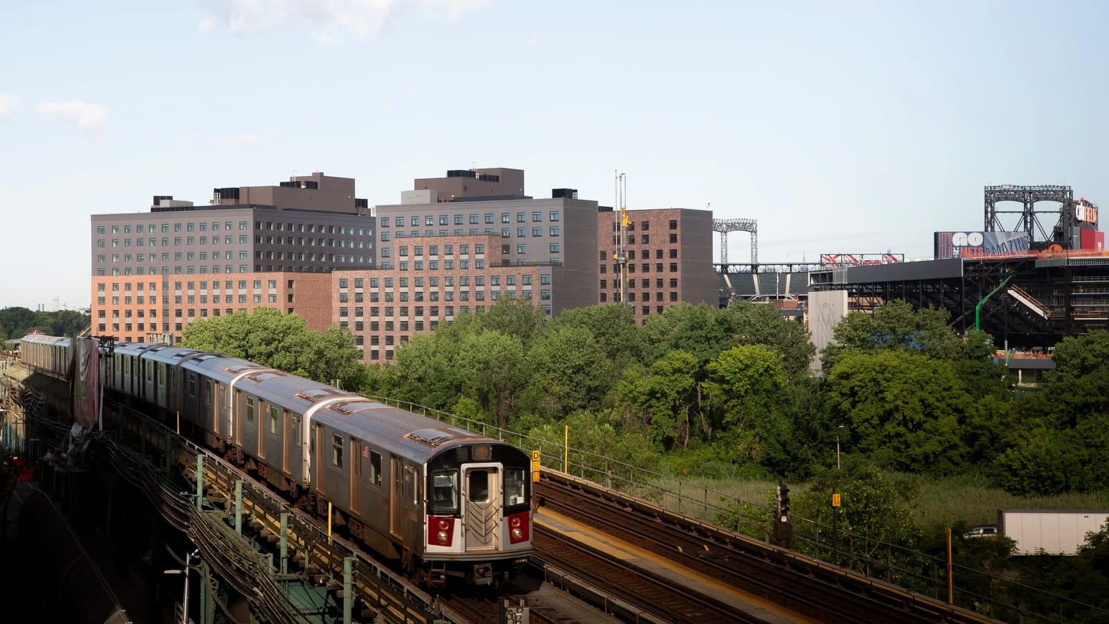 Photograph of a silver subway train on raised tracks in the foreground, with a group of mid-rise apartment buildings in red brick and grey panels beyond a band of trees, and stadium light towers, signs and a dark steel structure at the right