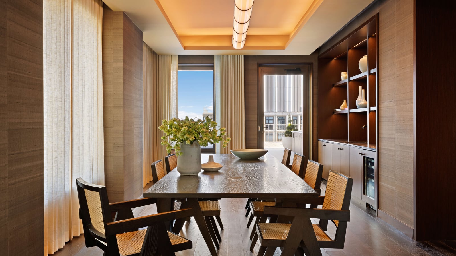 Photograph of a private dining room with a long dark wood table, cane-backed wooden chairs, grasscloth walls, a built-in cabinet with shelves of vases and a wine fridge, a long ceiling light, and a glass door opening onto a terrace