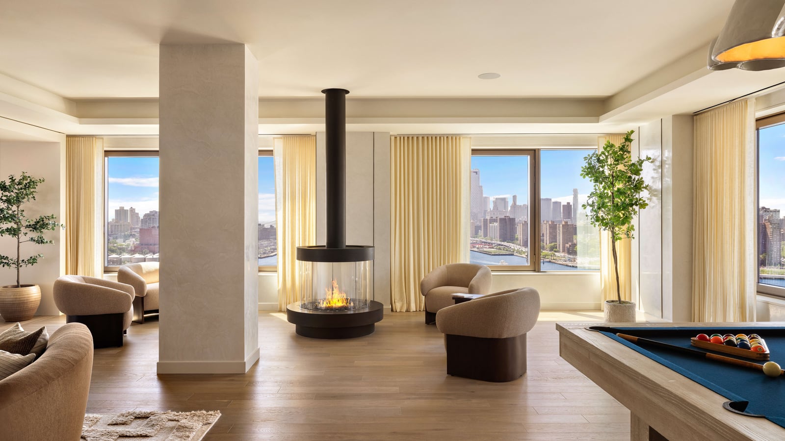 Photograph of a bright lounge with a round glass fireplace hanging from a black flue, curved beige armchairs, a pool table in the right foreground, potted trees and tall windows with cream curtains looking out over the East River and the Manhattan skyline