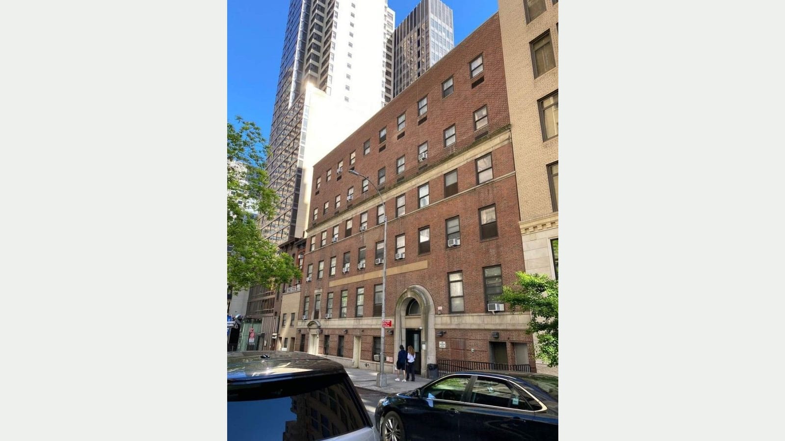 Photograph of a six-story red brick building with rows of small windows, air conditioners in some windows, a stone arched doorway with two people standing at it, a street lamp, parked black cars in front, a beige stone building at right and tall glass and white towers behind, under a blue sky