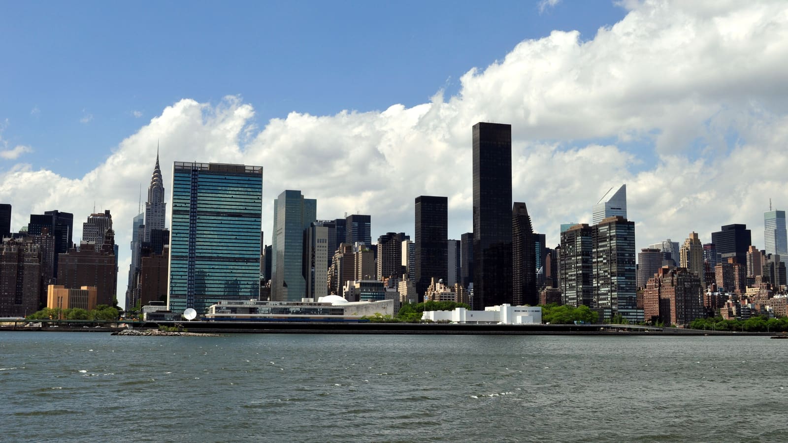 Photograph across a gray river of a skyline under blue sky and large white clouds: a tall green-glass slab, a low white building on the waterfront, a pointed silver spire at left and dark glass towers at center and right