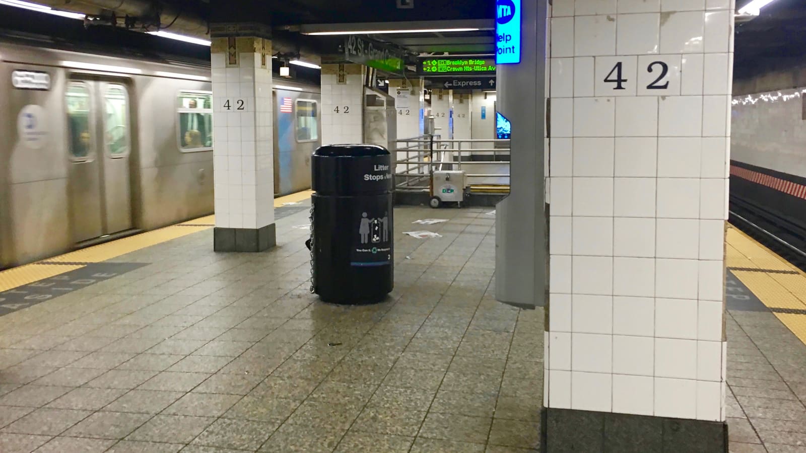 Photograph of an underground subway platform with white tiled columns marked 42, a black litter bin, a blue MTA Help Point sign, a green sign reading 42 St-Grand Central, an arrival board, and a silver train blurred in motion at left