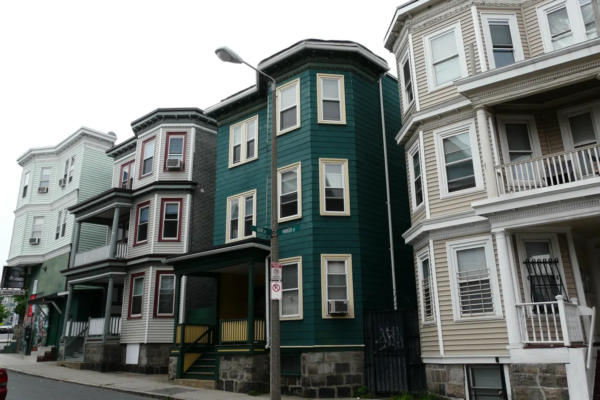 A row of three-story wooden triple-decker houses with bay fronts and porches, one painted dark green, at the corner of Gore Street and Parker Street, with a street lamp and parking signs