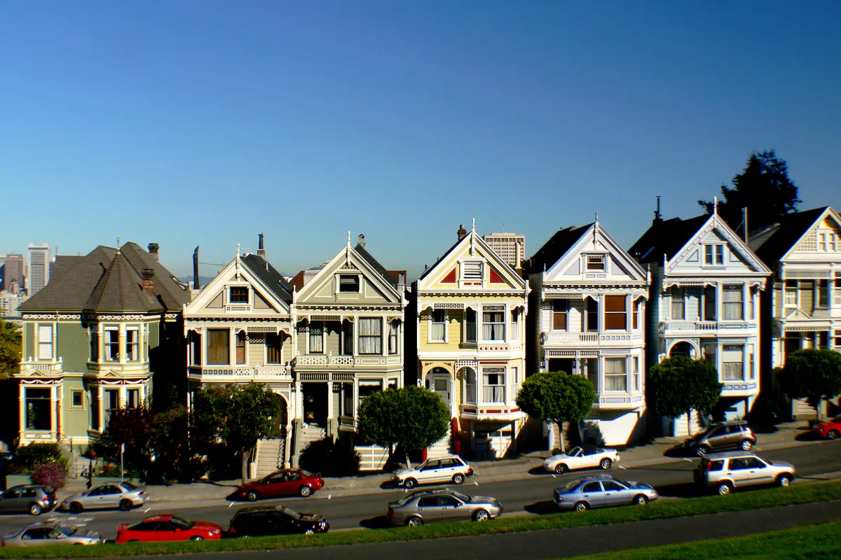Row of pastel Victorian houses with steep gables facing Alamo Square park in San Francisco, with parked cars along the street and downtown towers behind