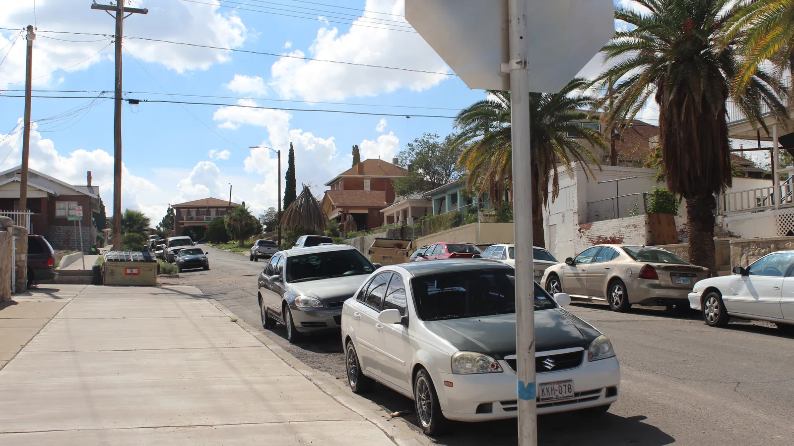 A sloping residential street in El Paso’s Sunset Heights neighborhood with palm trees, brick and stucco houses, and parked cars under a partly cloudy sky