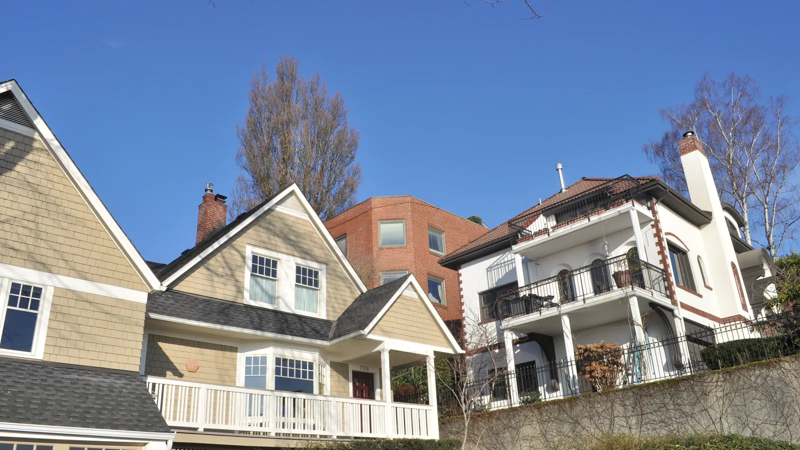 Shingled and stucco houses on a sloping street on southwest Queen Anne Hill in Seattle, with a brick apartment building behind them under a clear blue sky