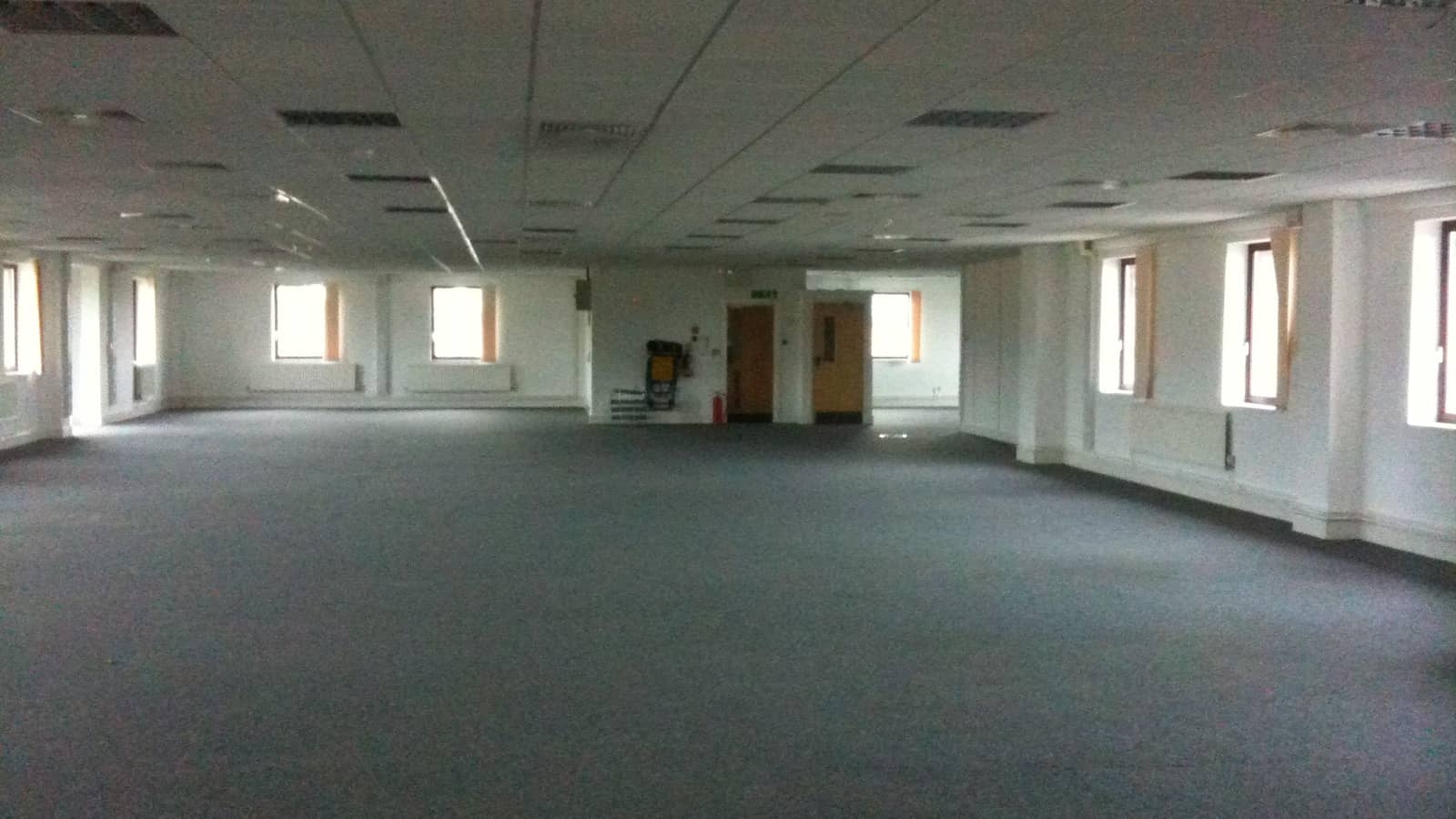 Empty office interior with gray carpet, white walls, perimeter windows and a suspended ceiling.