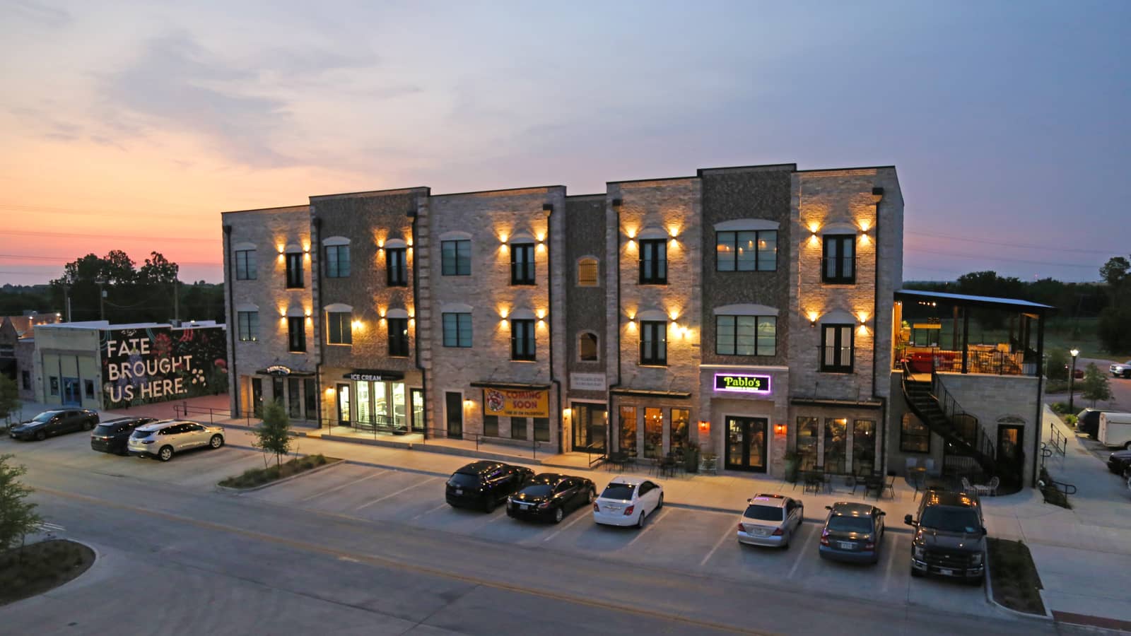 Three-story brick mixed-use building with ground-floor storefronts and upper windows at dusk in Fate, Texas.