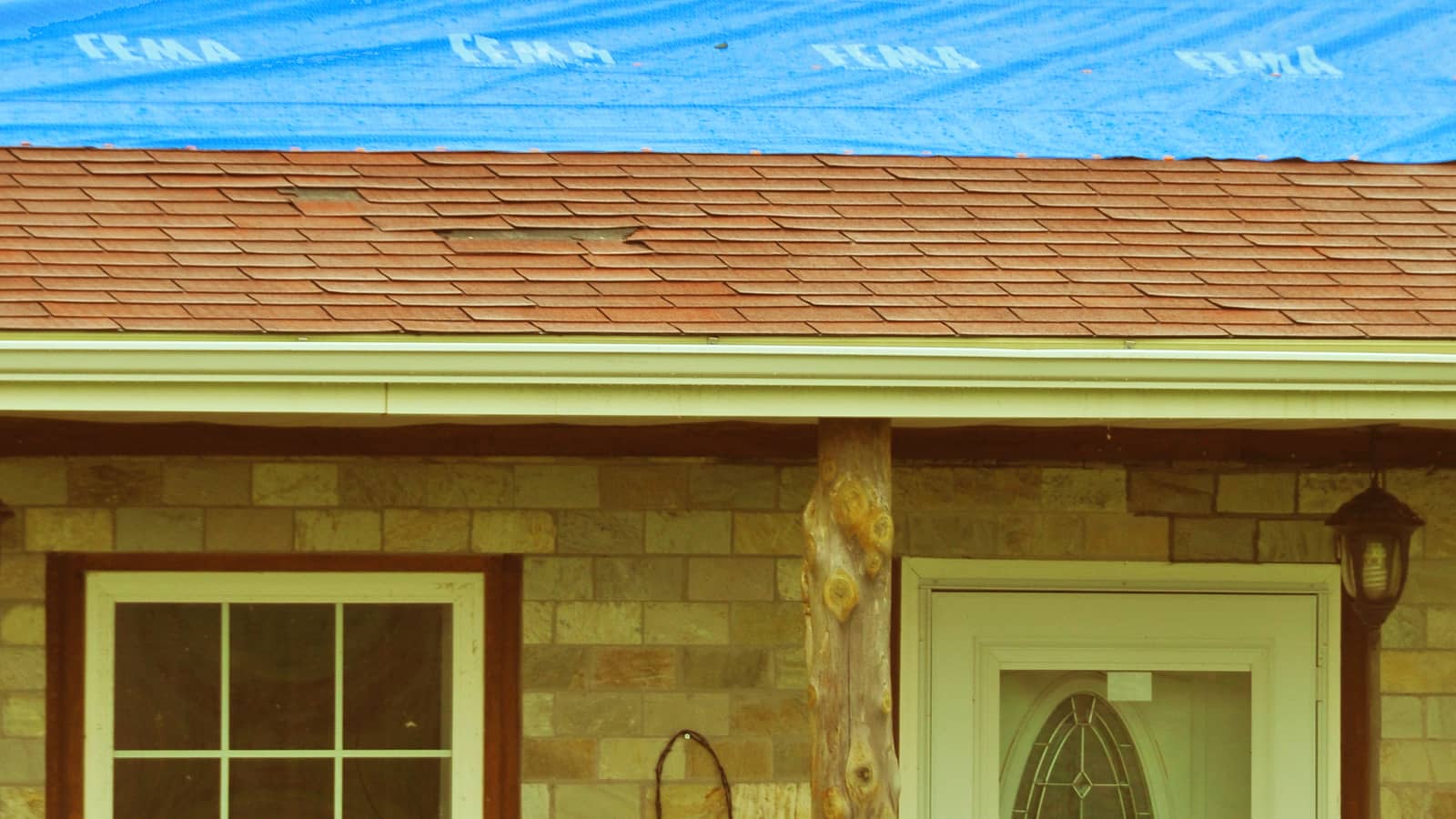 A blue tarp covers the upper part of a shingled roof above a stone-faced front porch.