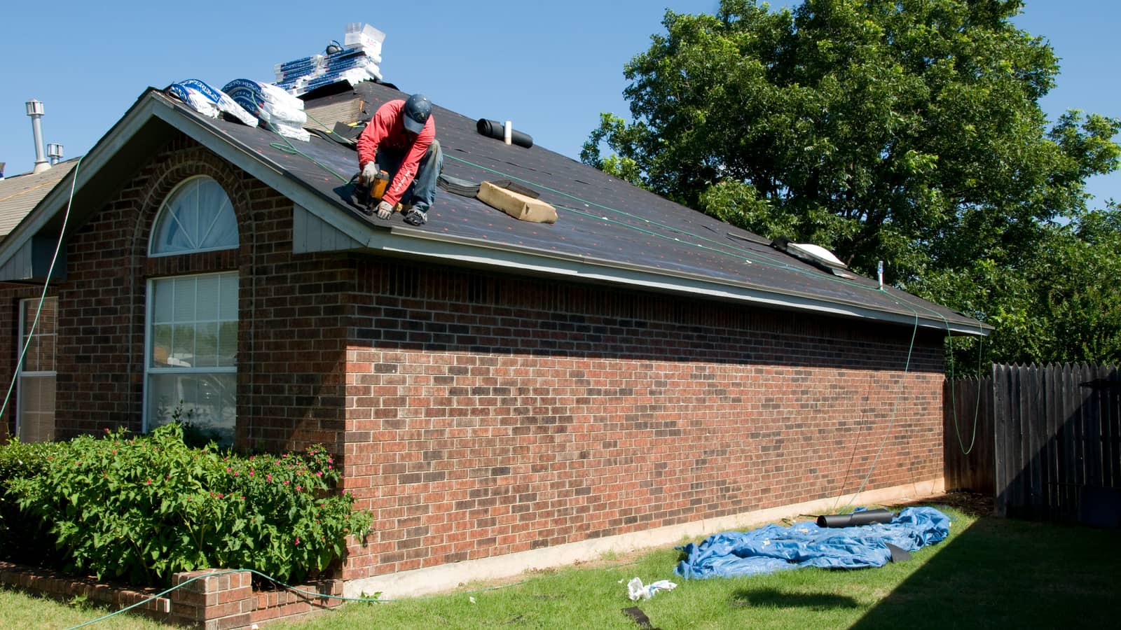 A roofer works on a sloping house roof with shingles and building materials beside him.