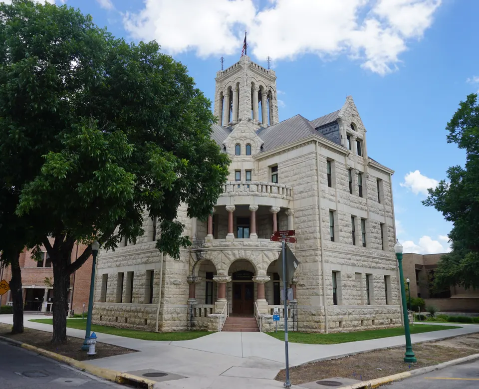 The limestone Comal County Courthouse with its central bell tower and arched porch in downtown New Braunfels, Texas, framed by large trees